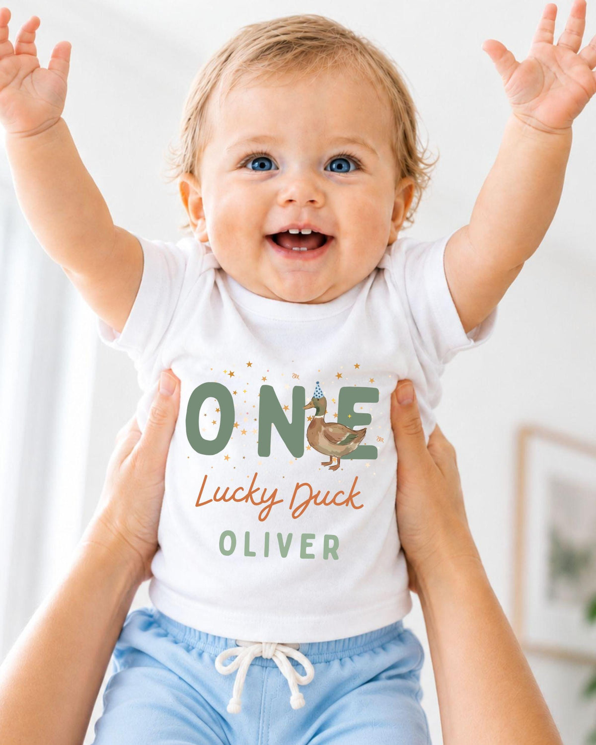 A smiling baby wearing a white shirt with the text "ONE Lucky Duck Oliver" is being held up by someone's arms.