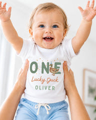 A smiling baby wearing a white shirt with the text "ONE Lucky Duck Oliver" is being held up by someone's arms.