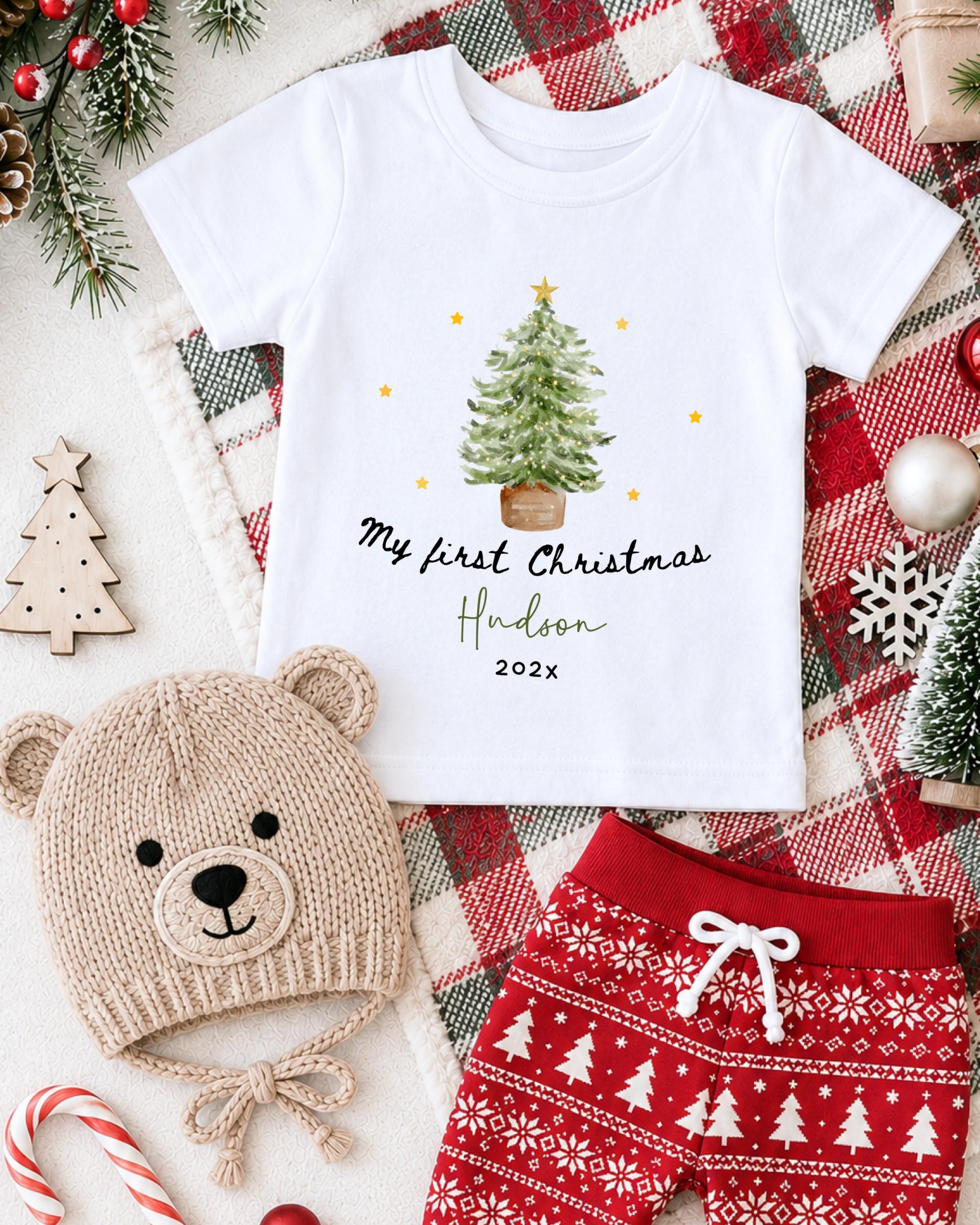 A white t-shirt with a Christmas tree design, a beige knit hat with a bear face, and red patterned shorts, all arranged on a plaid blanket with Christmas decorations.