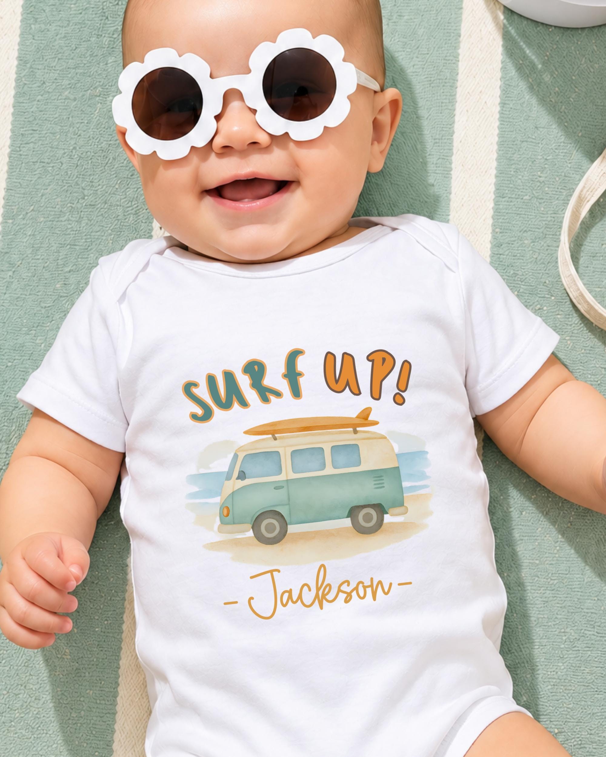 A baby wearing a white onesie with a surfboard graphic and sunglasses, smiling and laying on a green and white striped surface.