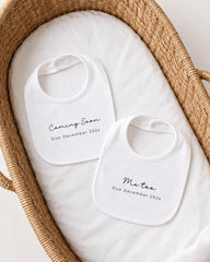 two white bibs with black text, placed in a woven basket. The text on the bibs announces the upcoming arrival of a baby, with the due date of December 2023.
