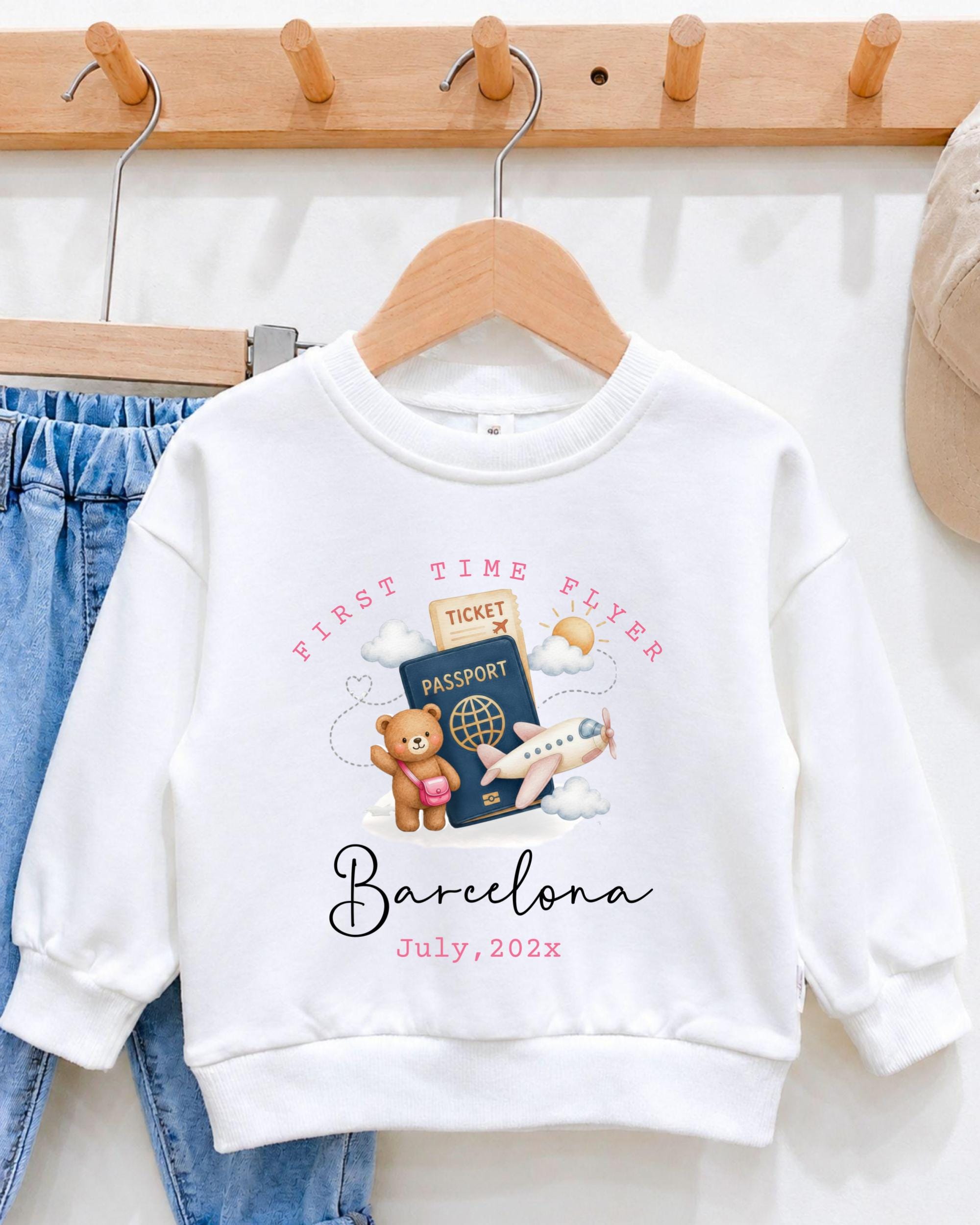 A white sweatshirt with a graphic design featuring a teddy bear, a passport, and an airplane, along with the text "First Time Flyer Barcelona July, 2023".