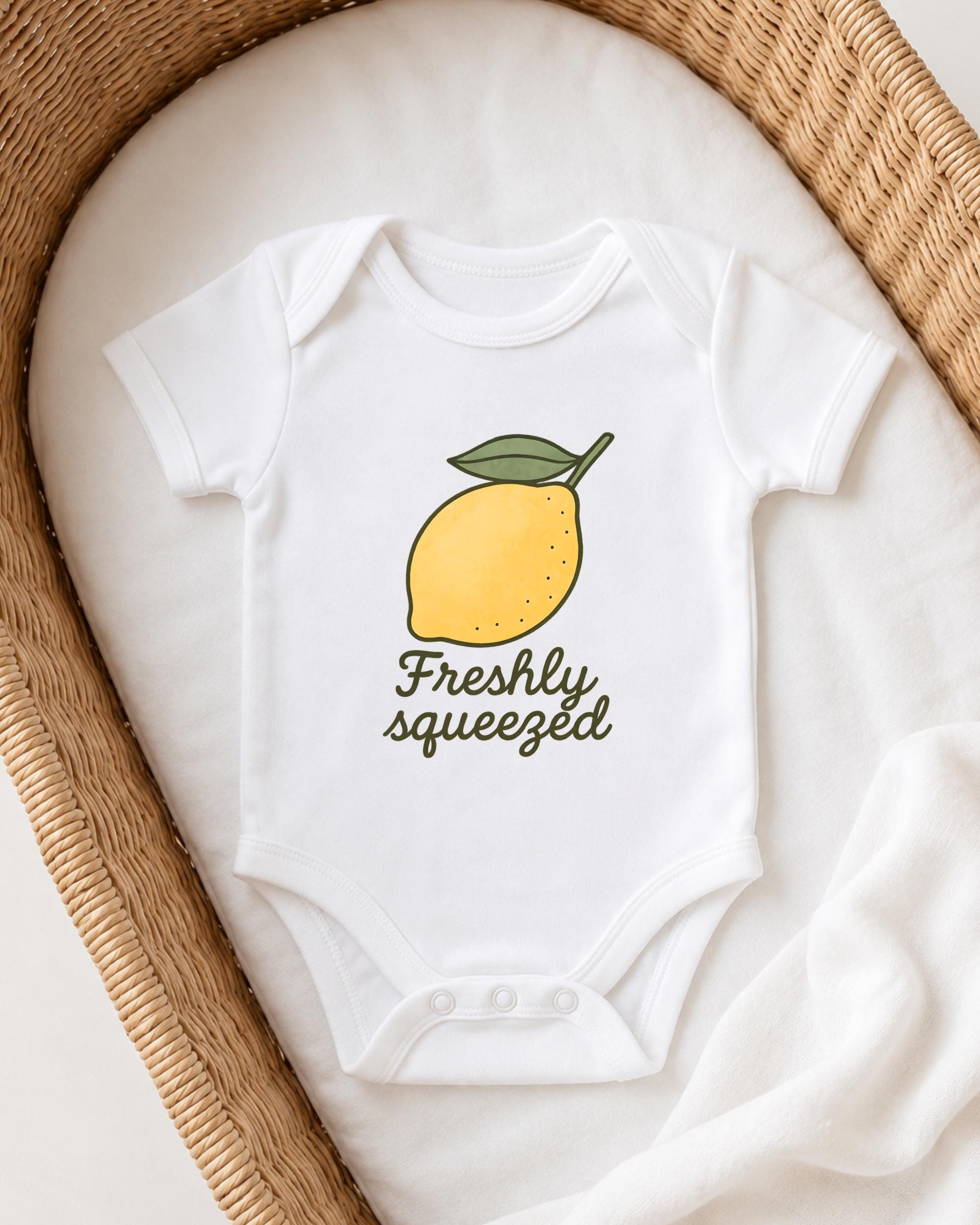 A white onesie with a lemon graphic and the text "Freshly squeezed" printed on it, placed on a white blanket inside a woven basket.