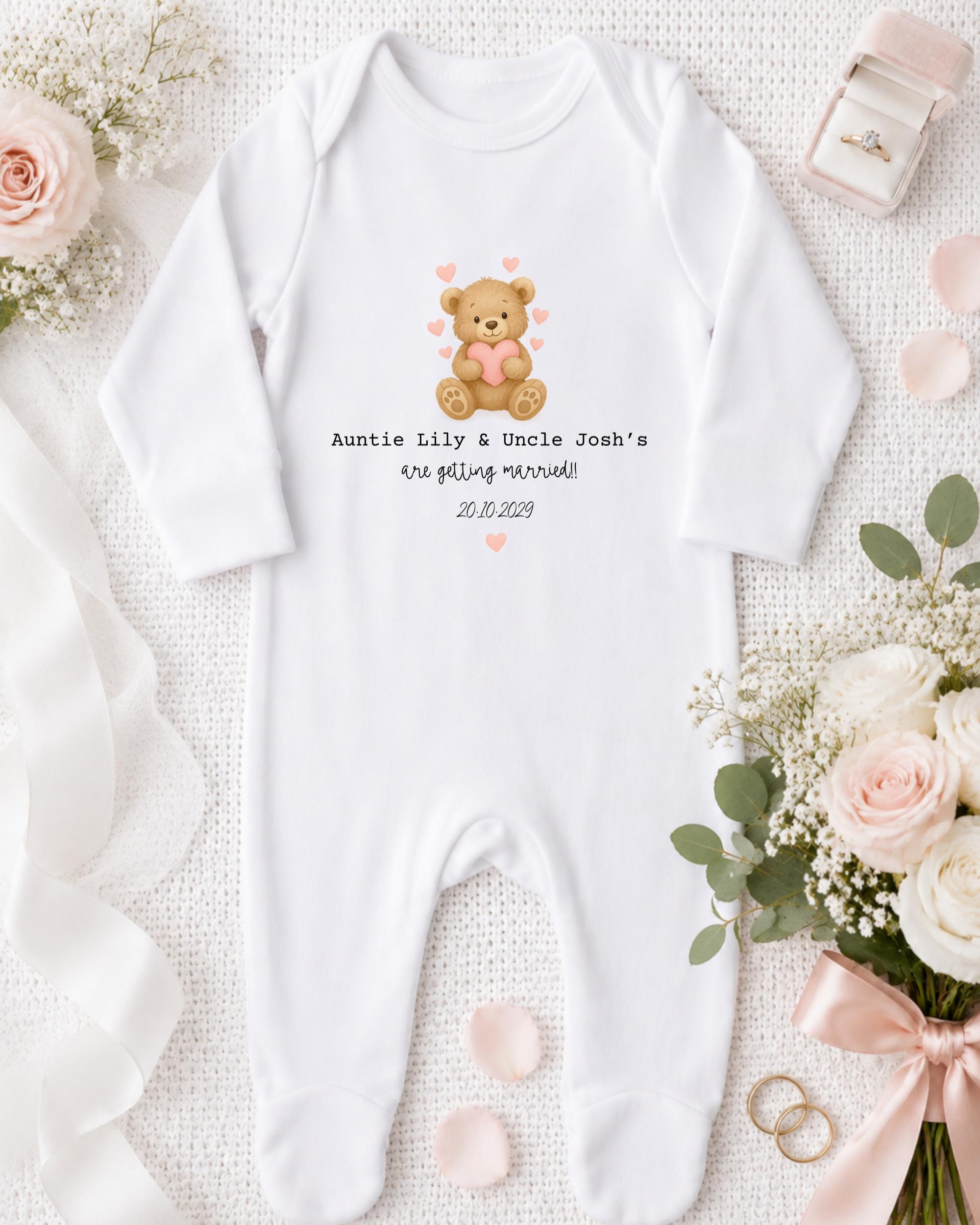 A white onesie with a teddy bear graphic and text, along with a pink ribbon, a ring, and some flowers, all arranged on a white background.