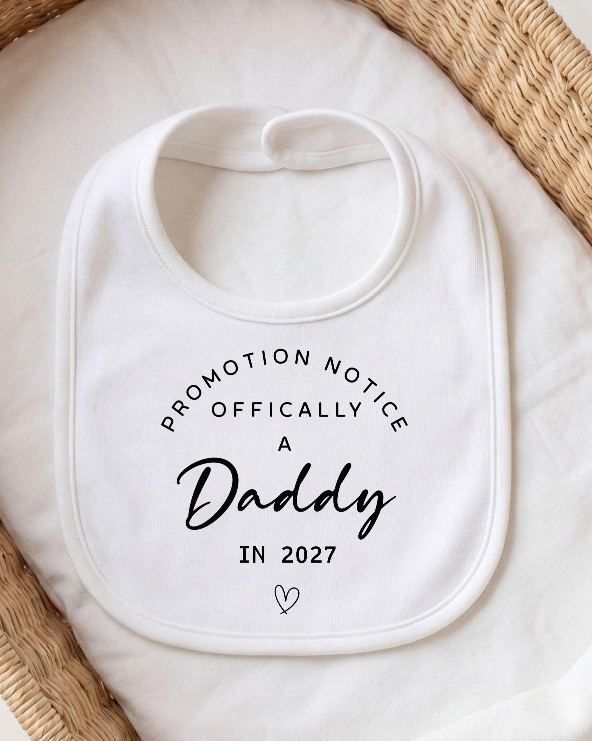 A white bib with black text that reads "Promotion Notice Officially A Daddy in 2027"