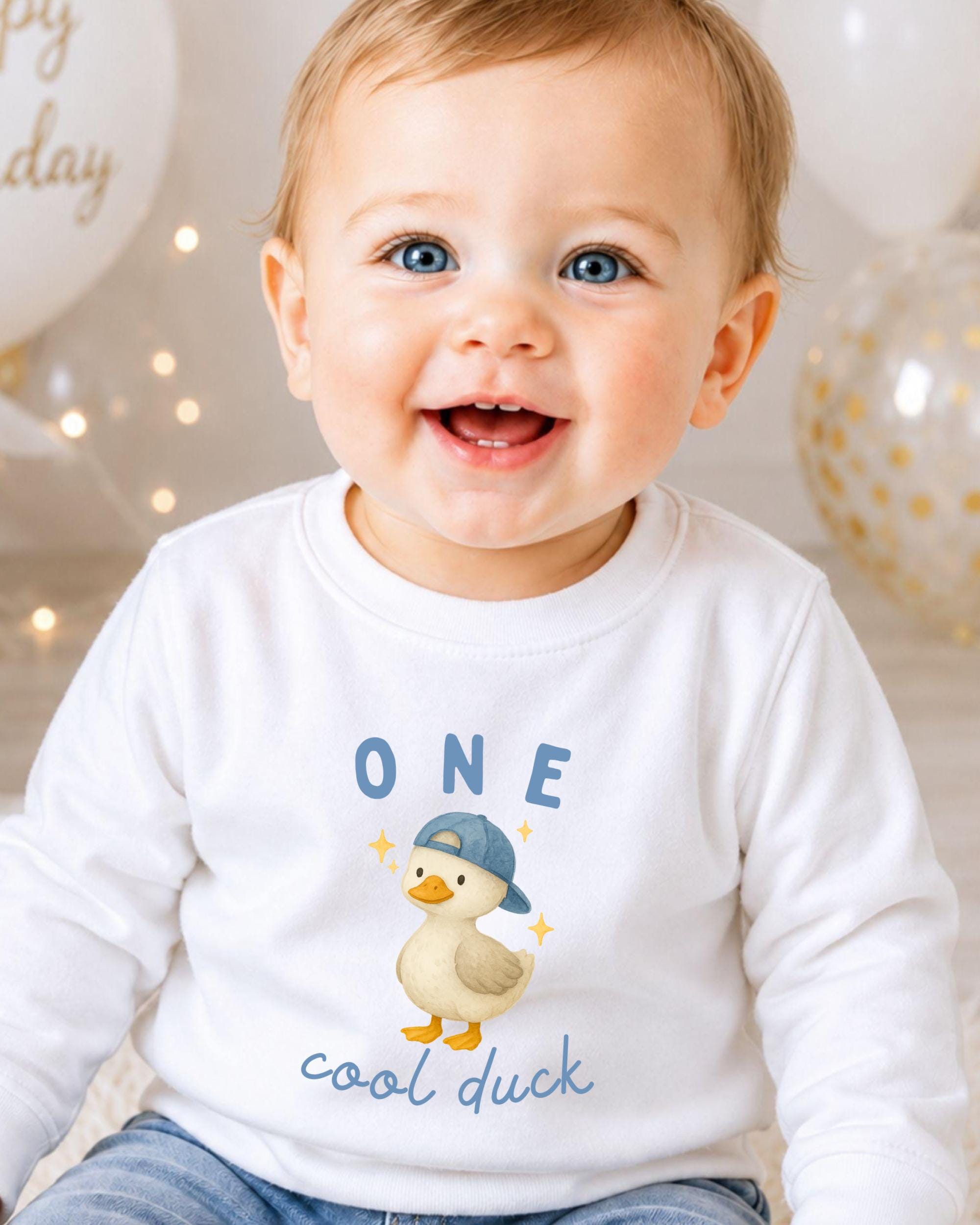 A smiling baby boy wearing a white shirt with a cartoon duck and the text "One cool duck" is the focus of the image.