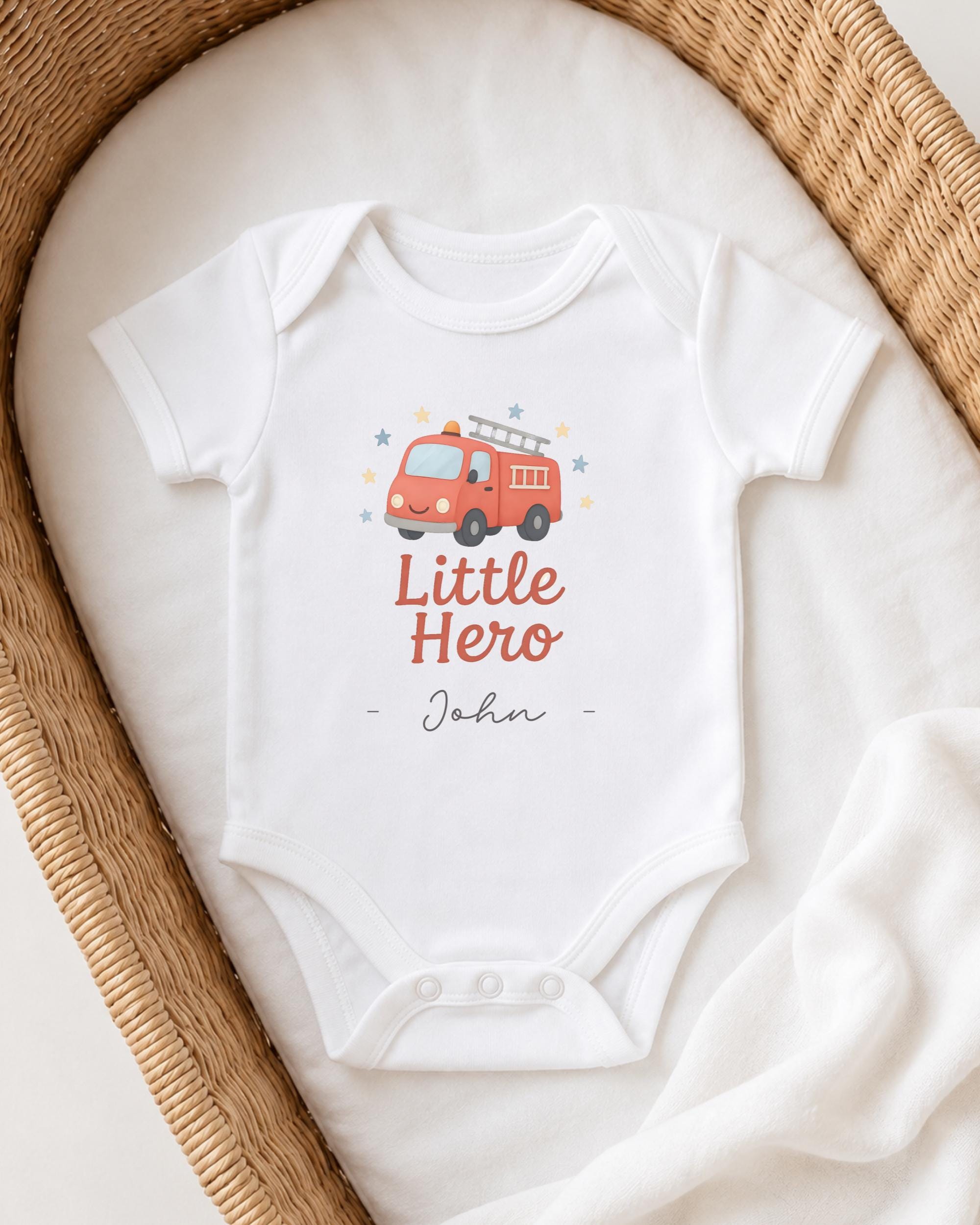 A white baby onesie with a fire truck graphic and the text "Little Hero John" printed on it, placed in a woven basket.