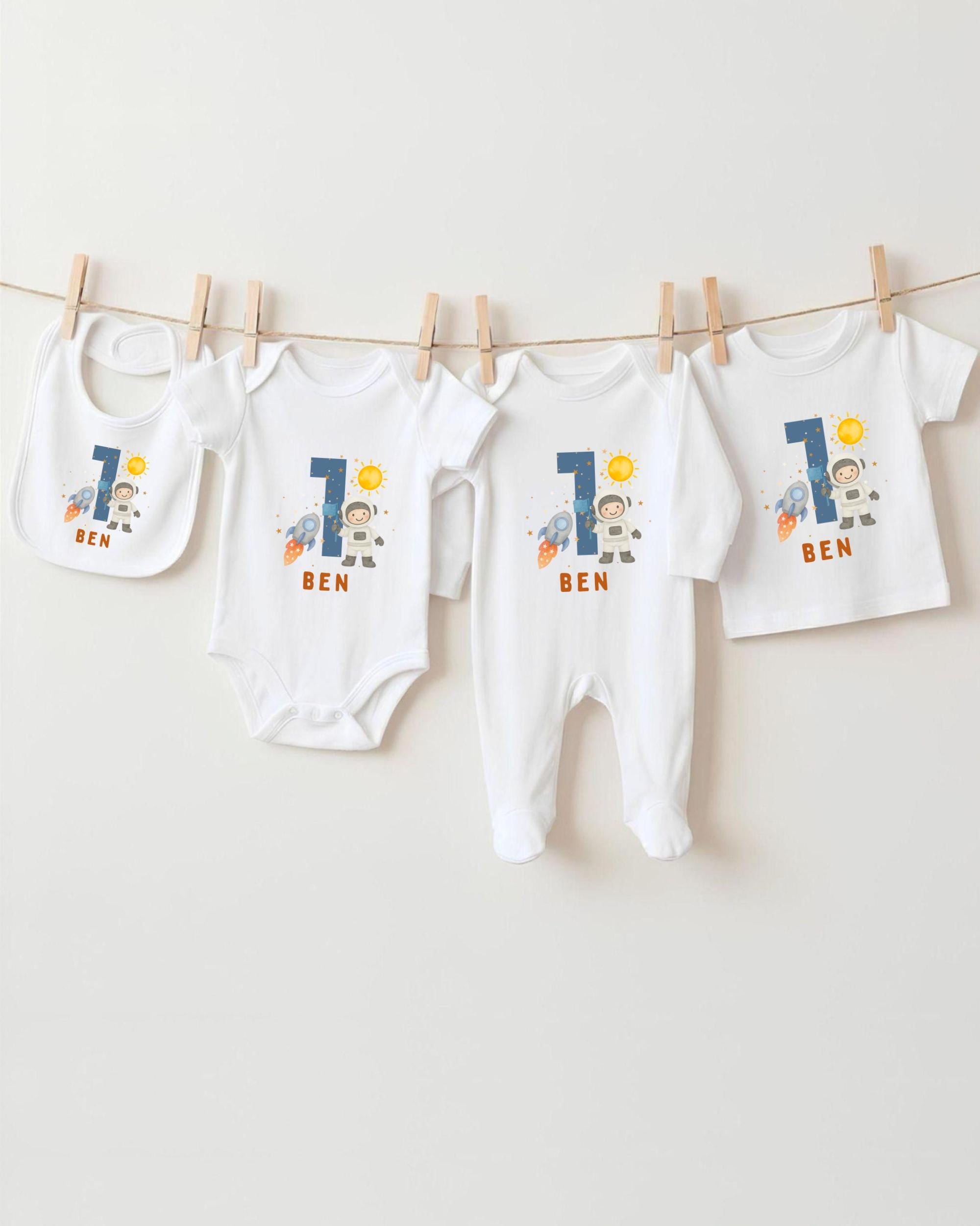 A collection of white baby clothes, including onesies and bibs, with the name "Ben" printed on them. The clothes are hanging on a clothesline, and there are four of them in total.