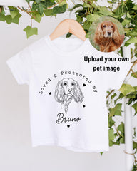 A white t-shirt with a graphic design featuring a dog's face and the text "Loved & Protected by Bruno" printed on it.