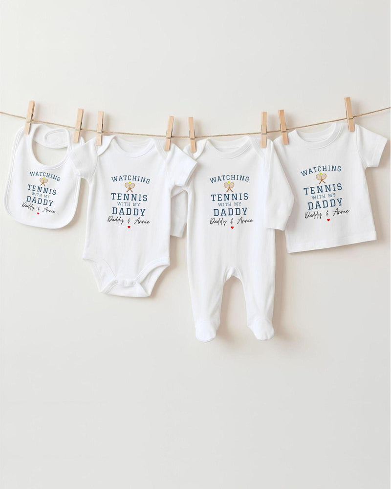 A row of four white baby clothes with the text "WATCHING TENNIS DADDY Daddy & Ava" printed on them, hanging on a clothesline.