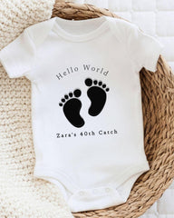 A white baby onesie with a black footprint design and text printed on it, placed on a woven basket.
