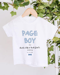 A white t-shirt with the text "PAGE BOY" printed on it, along with a date and a name. The t-shirt is hanging on a wooden hanger, and there are green leaves visible in the background.