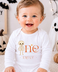 A smiling baby boy wearing a white onesie with a skeleton and stars on it, sitting in front of Halloween decorations.