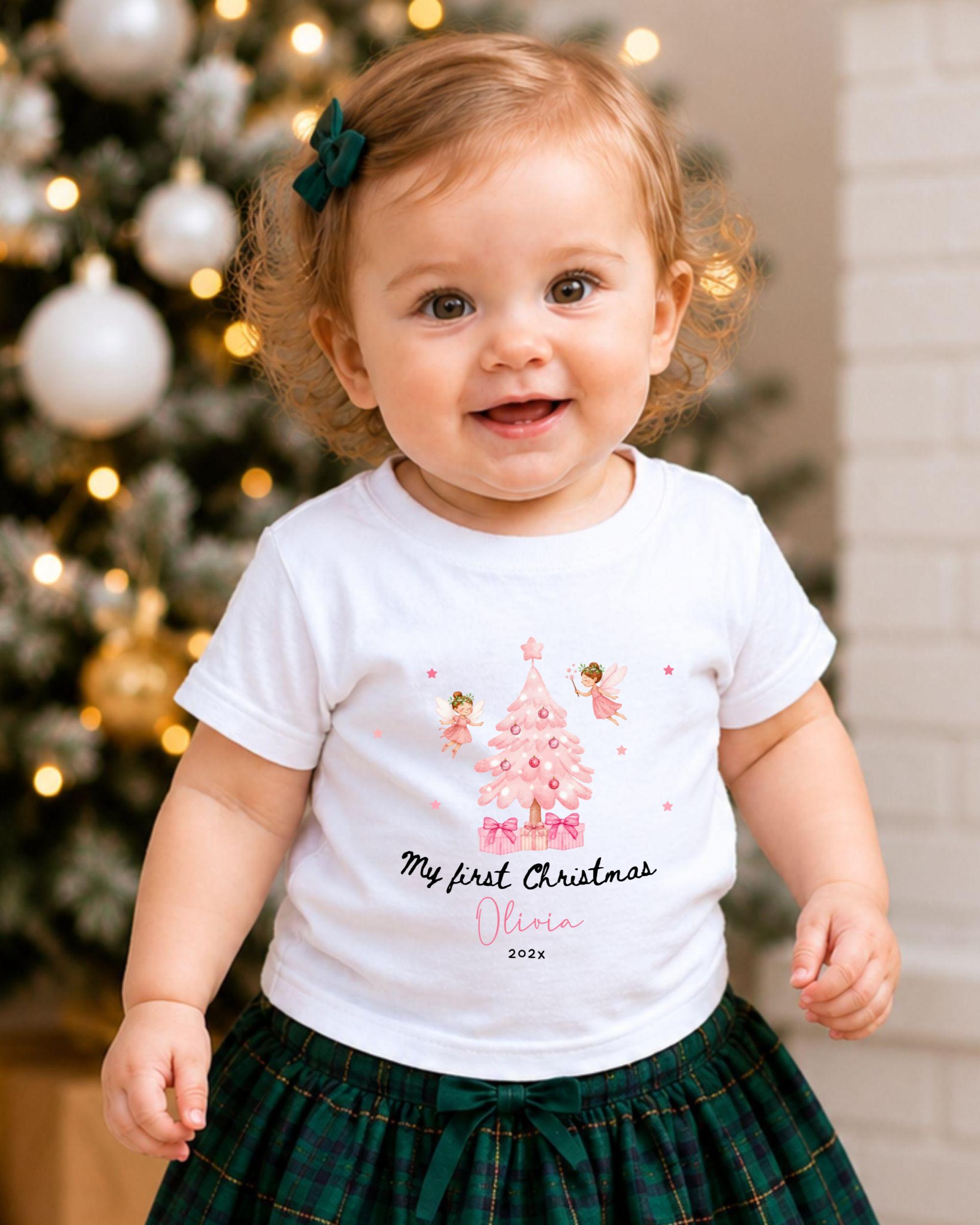 A smiling baby girl wearing a white t-shirt with a Christmas tree design and text, standing in front of a Christmas tree with lights.