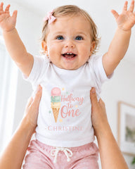 A smiling baby girl wearing a white t-shirt with a personalized text and an ice cream cone graphic, being held up by someone's arms.