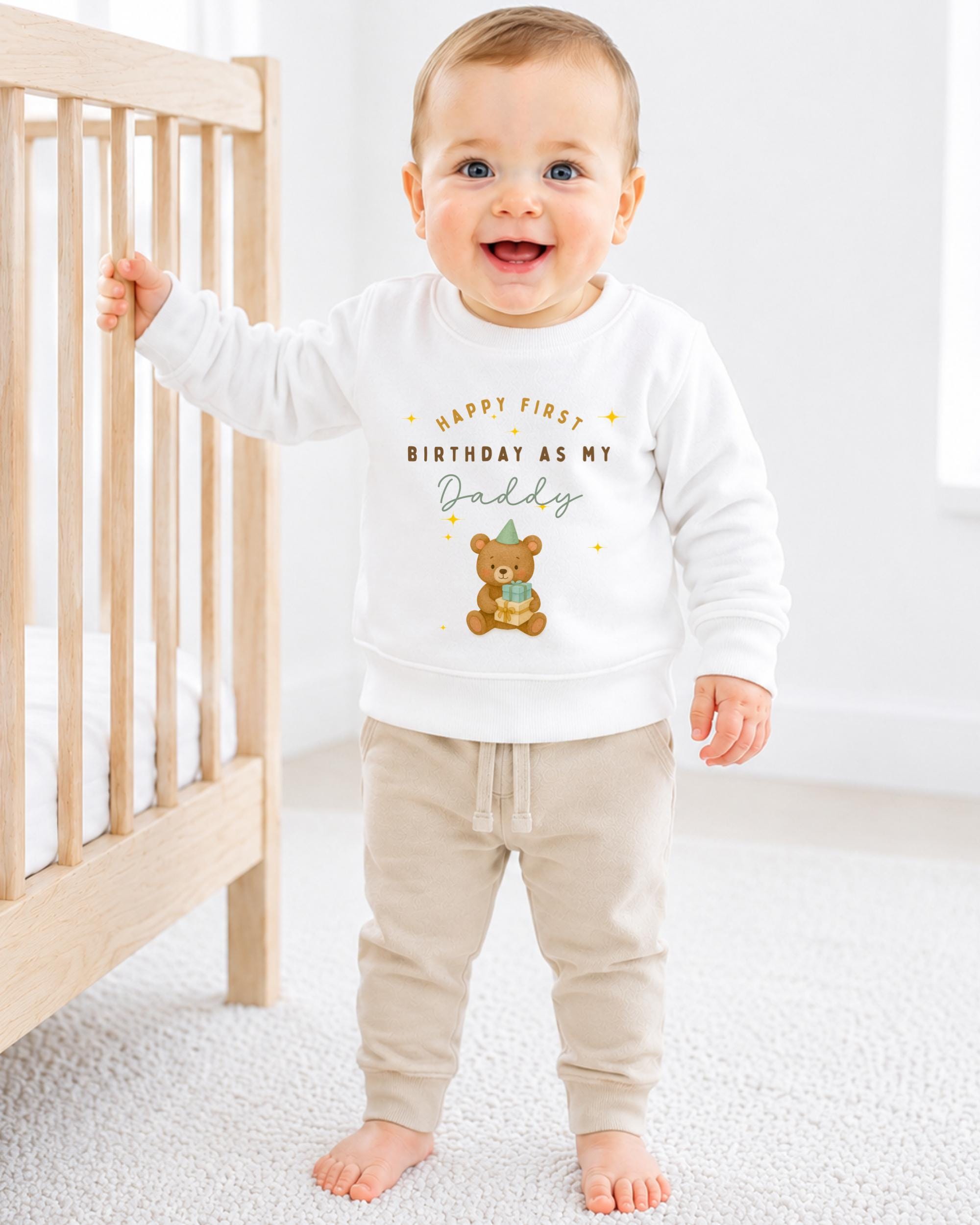 A smiling baby boy wearing a white sweatshirt with a teddy bear and text that says "Happy first birthday as my Daddy".