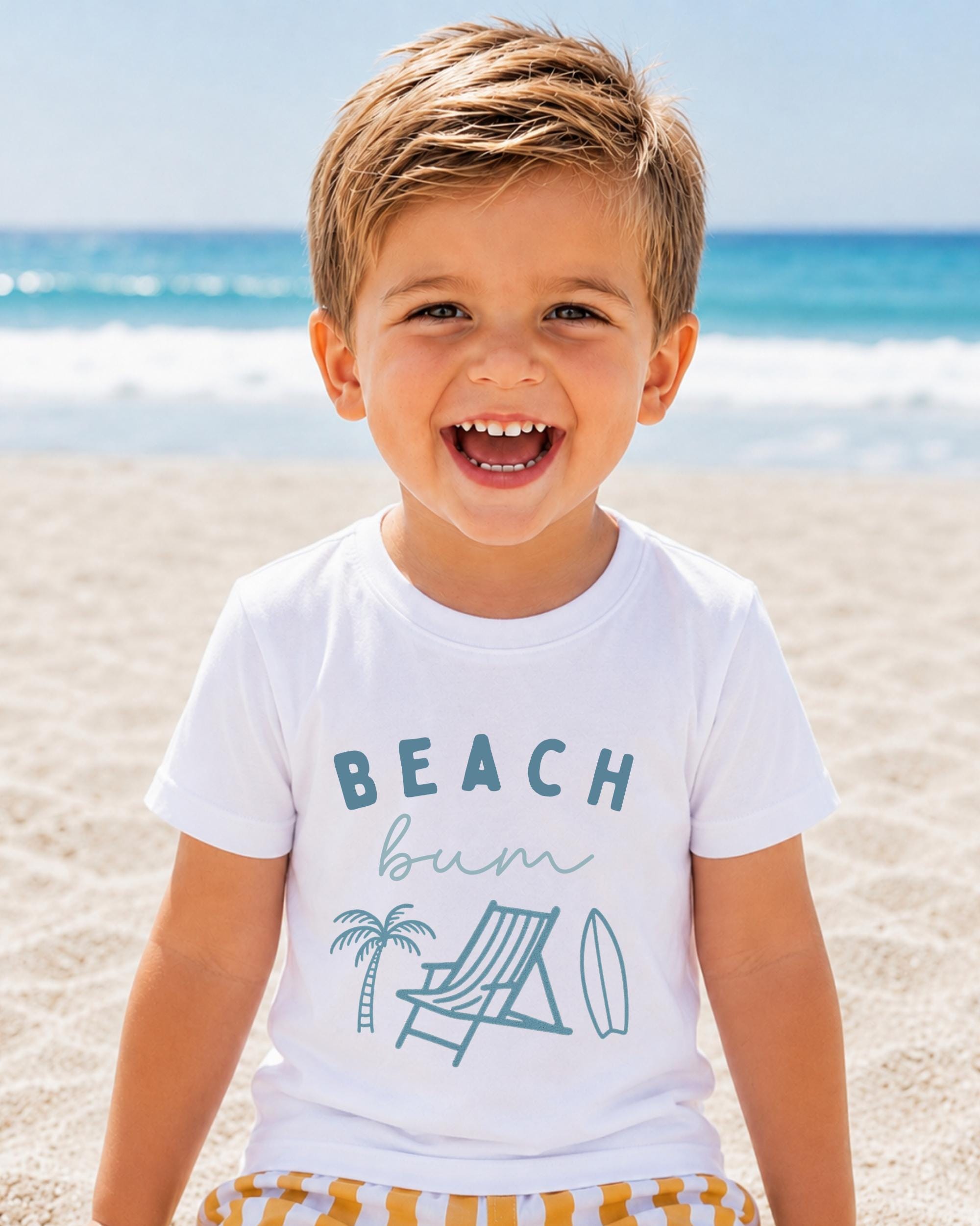 A young boy with a big smile on his face, wearing a white t-shirt with a beach-themed graphic and text, is standing on a sandy beach with the ocean in the background.