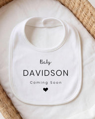 A white bib with the text "Baby Davidson Coming Soon" printed on it, placed on a white cloth.