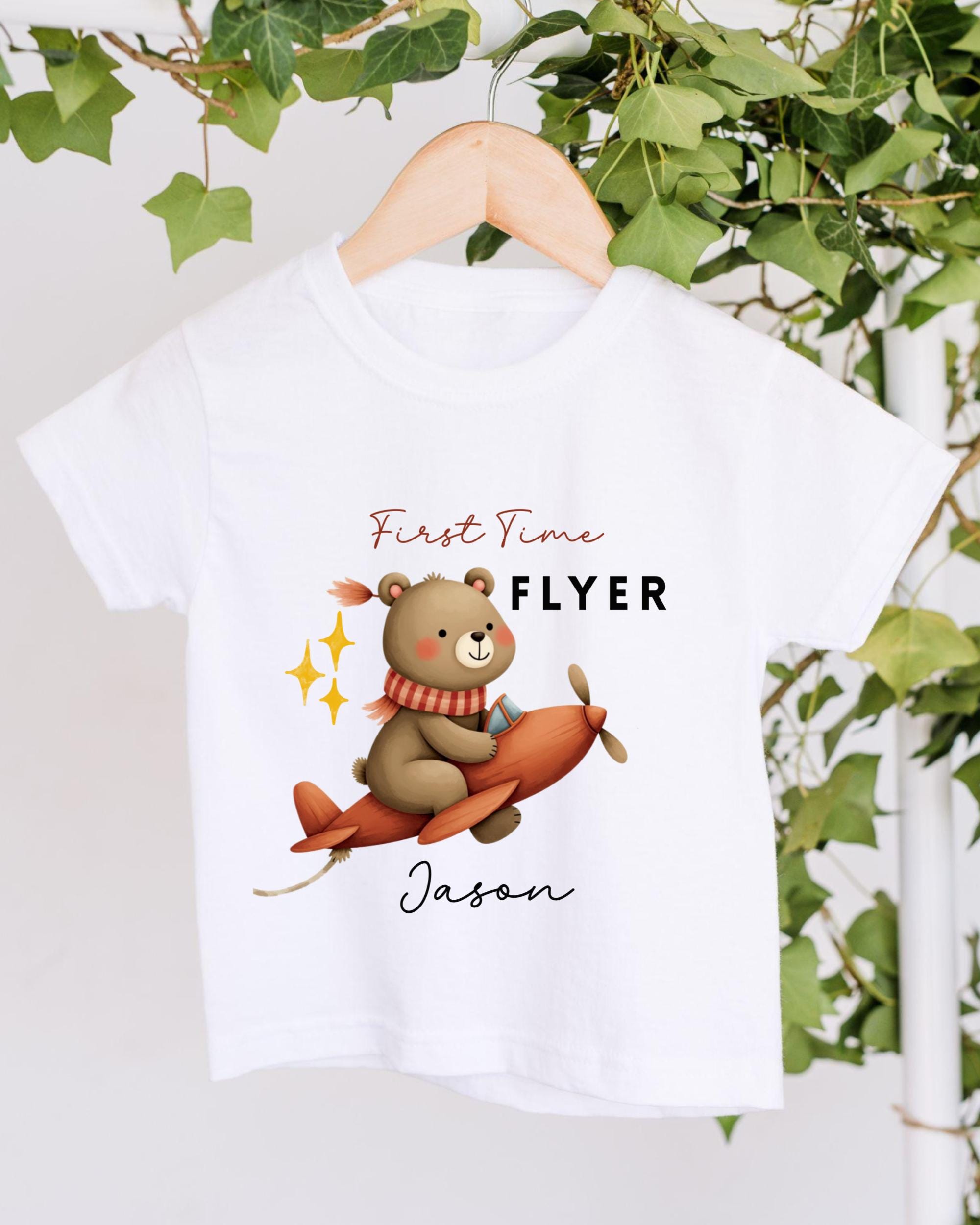 A white t-shirt with a cartoon image of a bear sitting on a plane and the text "First Time Flyer Jason" printed on it.