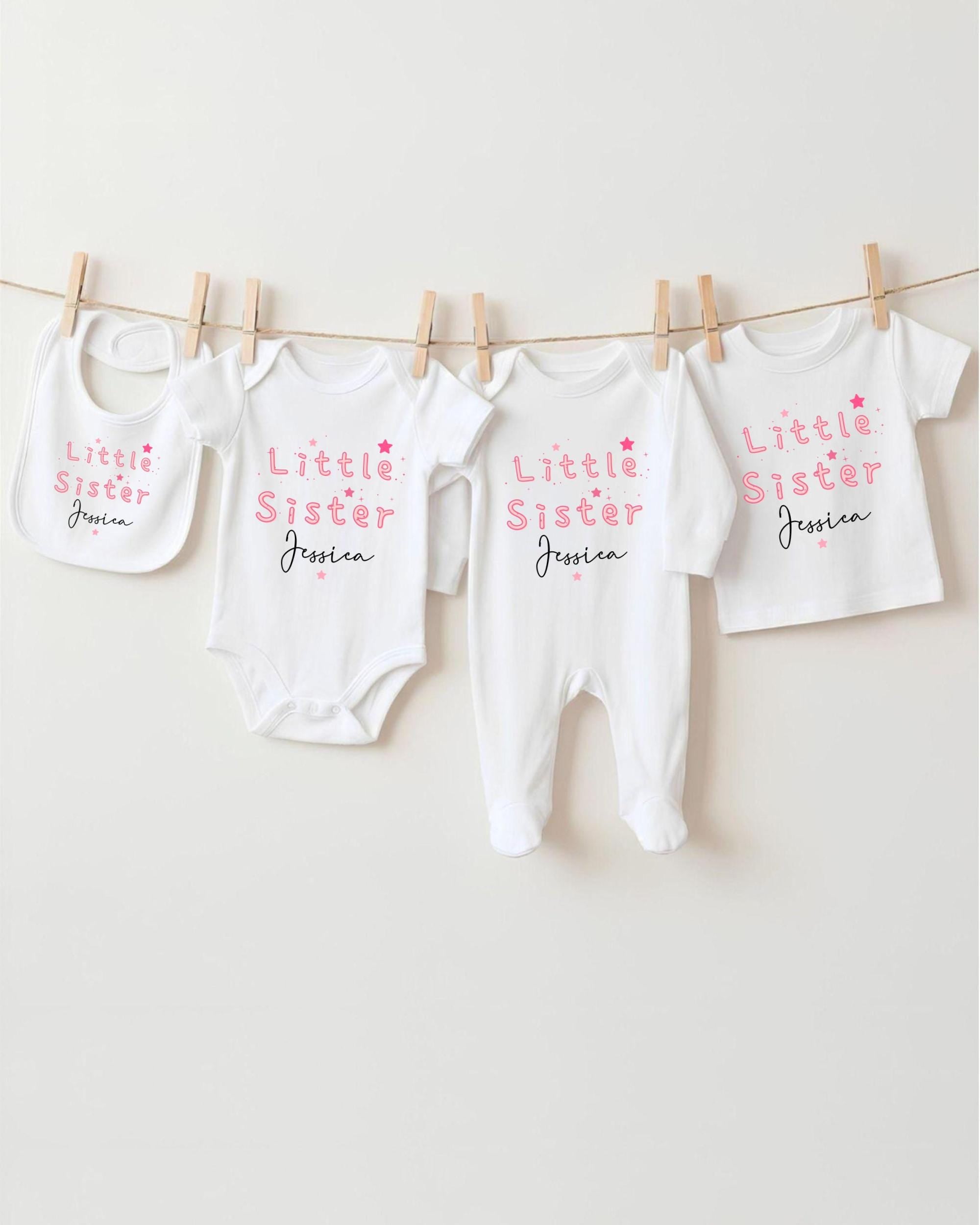 A row of four white baby clothes with the text "Little Sister" printed on them, hanging on a clothesline.
