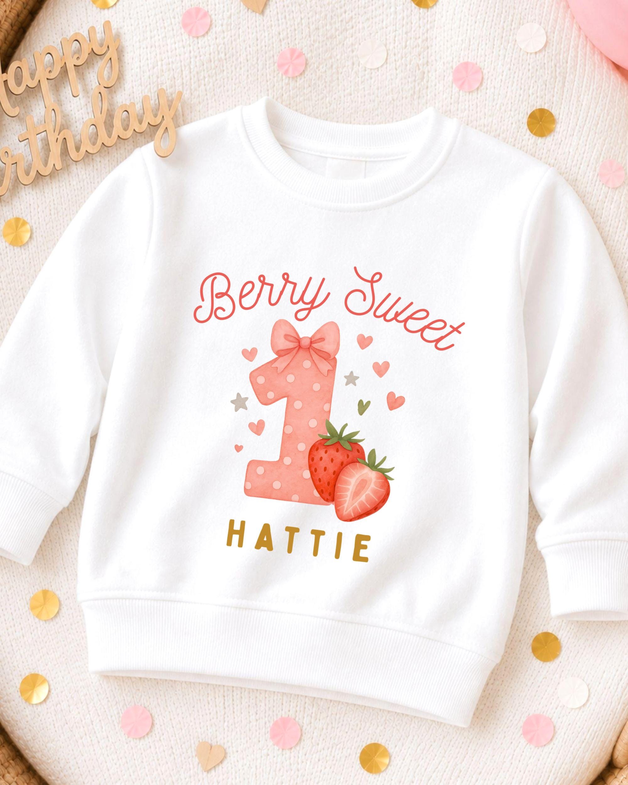 A white sweatshirt with a personalized design for a first birthday, featuring a pink polka dot number one, strawberries, and the text "Berry Sweet" and "Hattie".