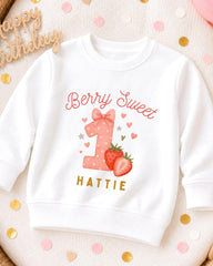 A white sweatshirt with a personalized design for a first birthday, featuring a pink polka dot number one, strawberries, and the text "Berry Sweet" and "Hattie".