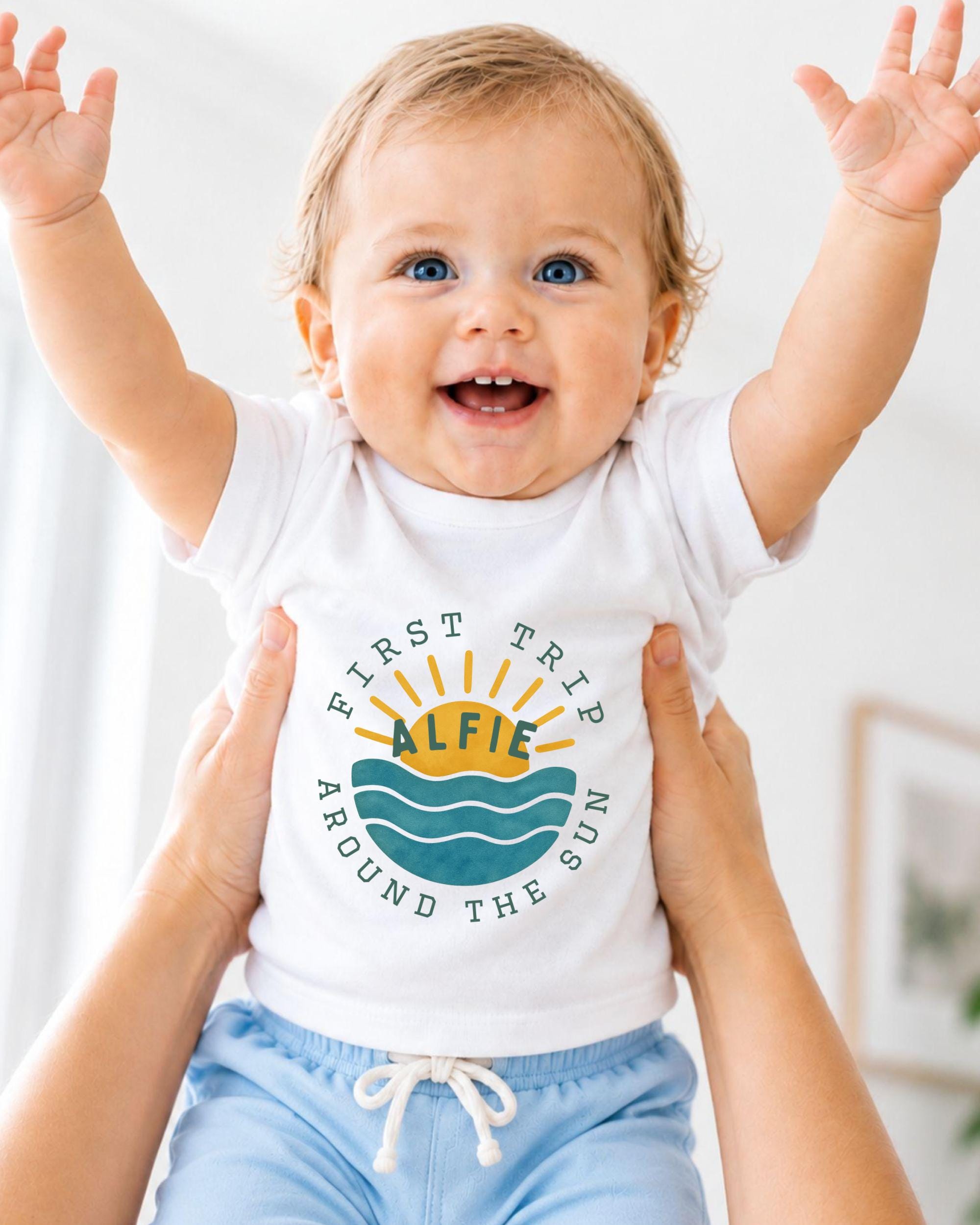 A smiling baby wearing a white shirt with a graphic design and text, being held up by someone's arms.