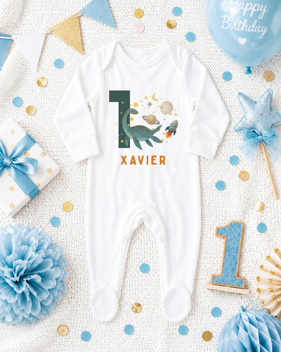 A white bodysuit with the number 10 and the name "XAVIER" printed on it, surrounded by various baby-themed items such as a blue star, a blue ribbon, and confetti.
