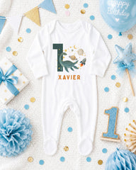 A white onesie with the number 10 and the name "XAVIER" printed on it, surrounded by various baby-themed items such as a blue star, a blue ribbon, and confetti.