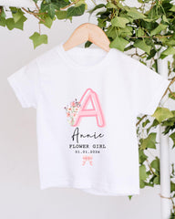 A white t-shirt with a pink floral design and the text "A Annie Flower Girl 01.01.2023" printed on it, hanging on a wooden hanger.