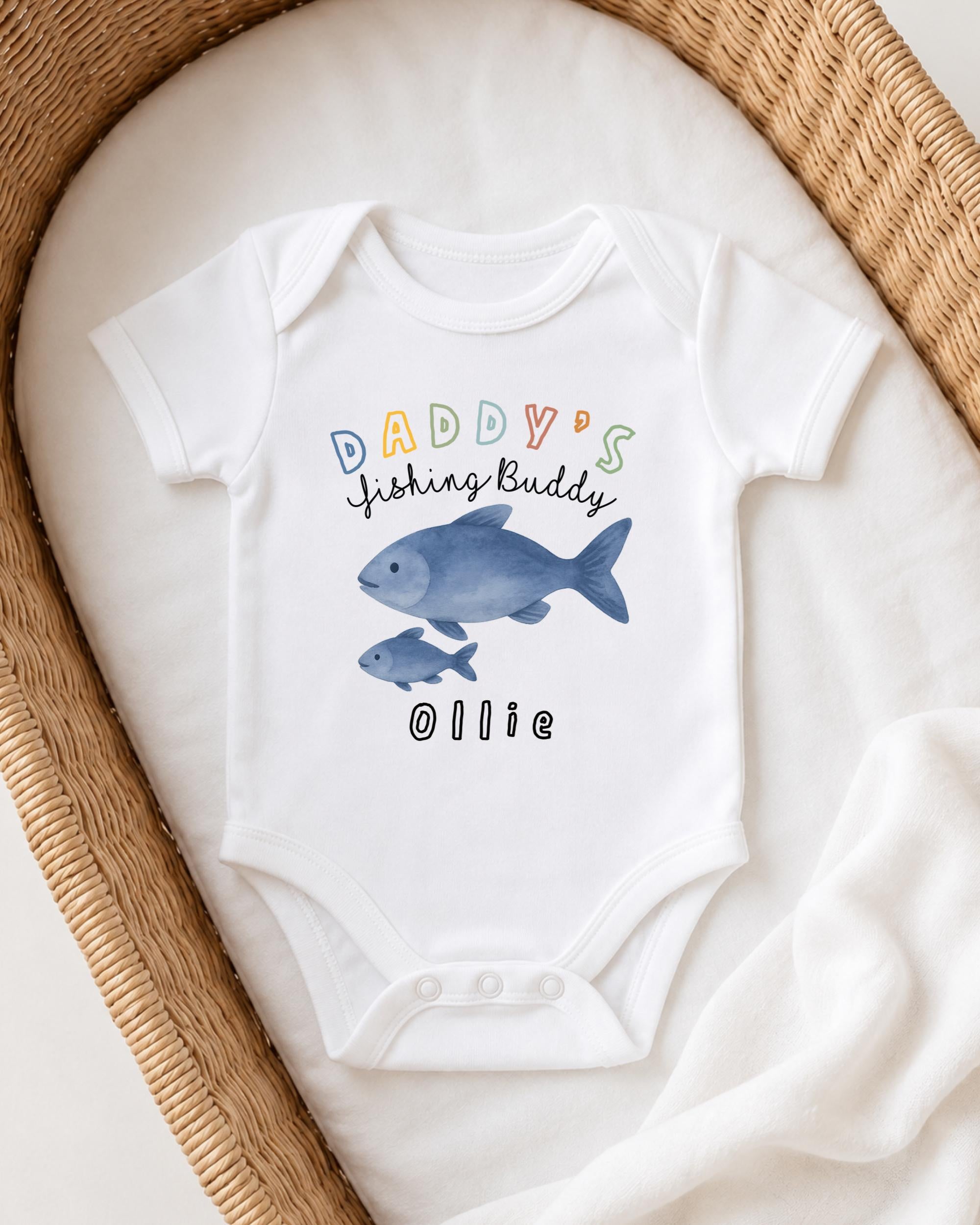 A white onesie with a fish design and the text "DADDY'S FISHING BUDDY OLLIE" printed on it, placed on a white blanket.