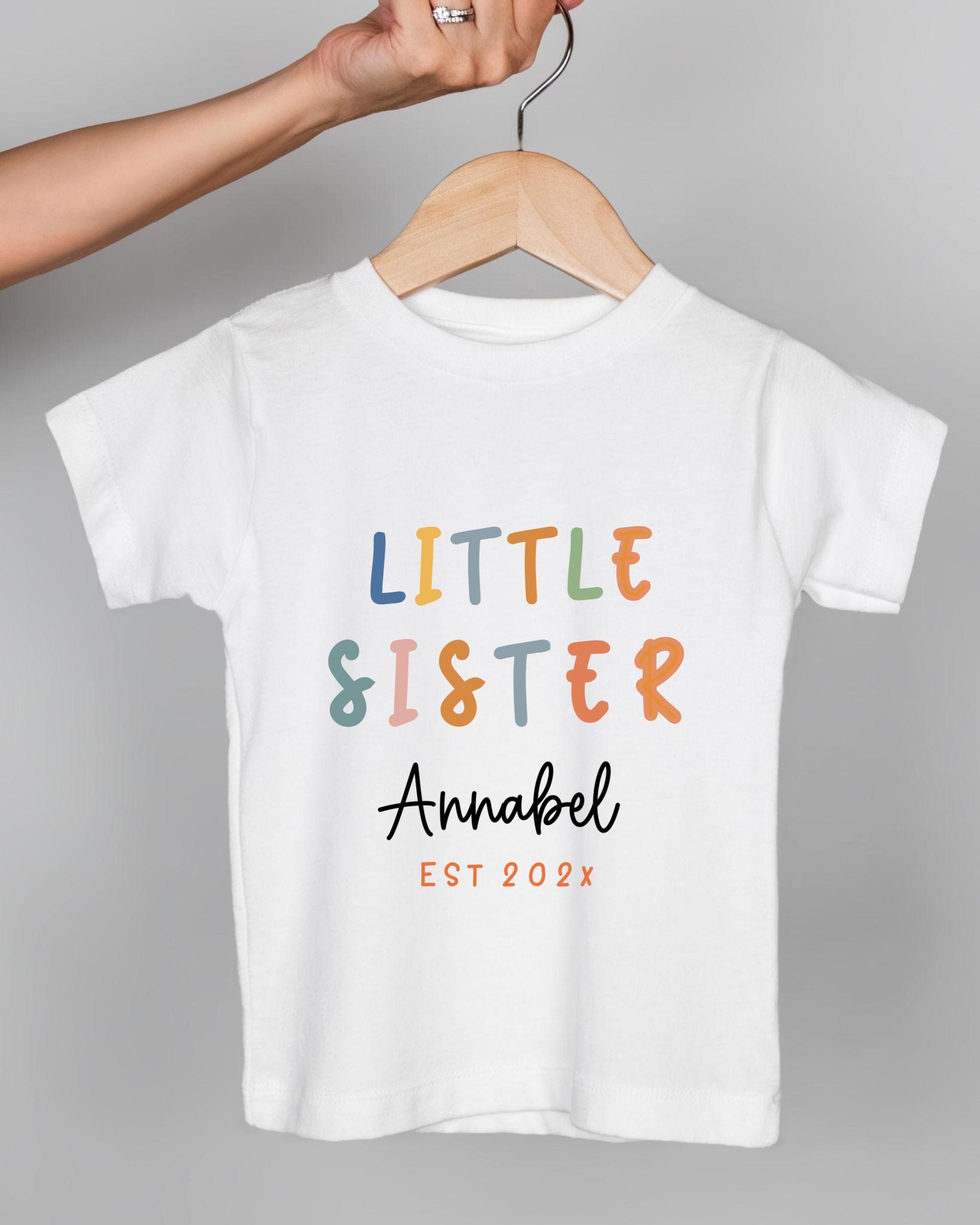 A white t-shirt with the text "LITTLE SISTER Annabel" printed on it, along with the year "EST 202x" below it.