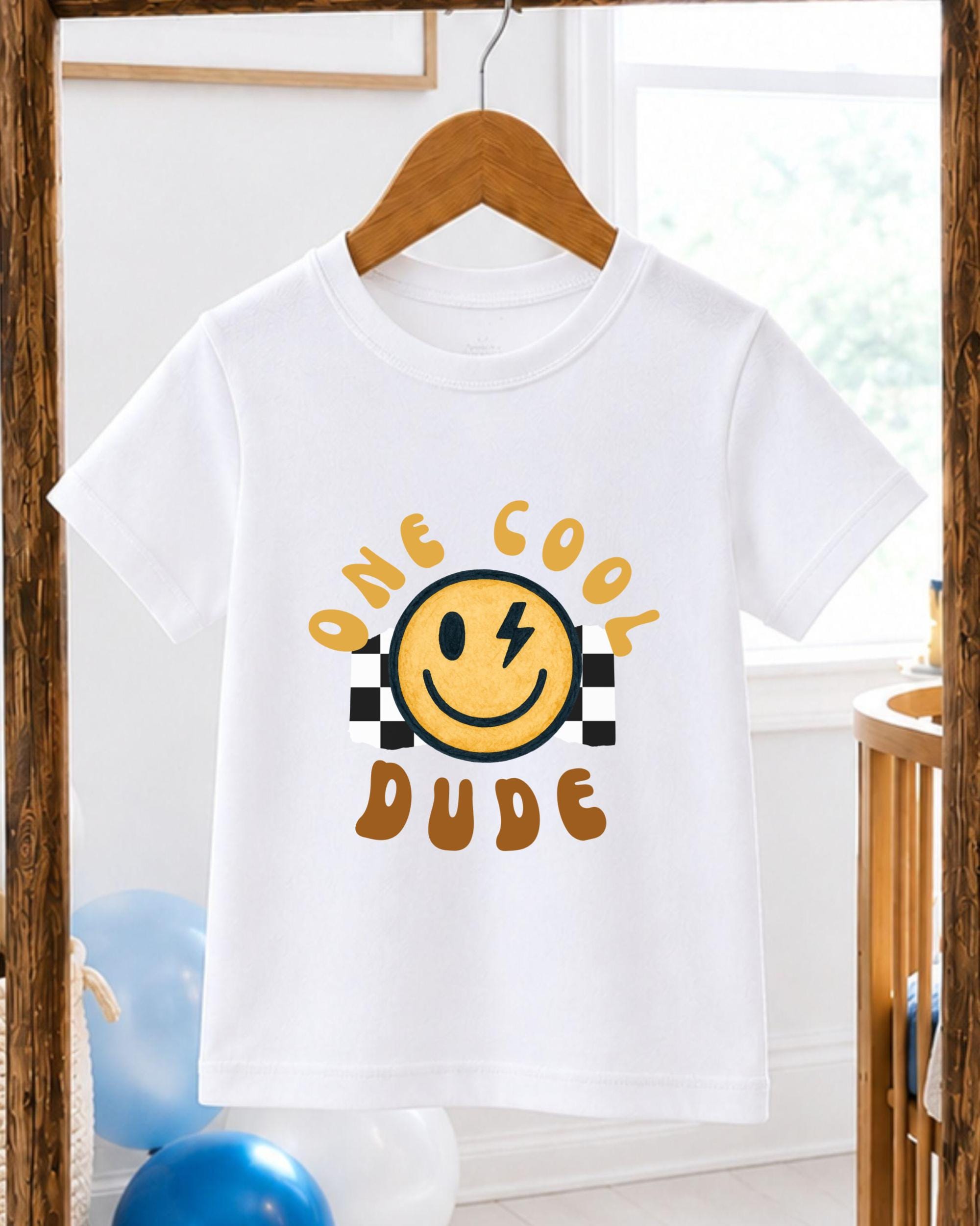 A white t-shirt with a yellow smiley face and the text "One cool dude" printed on it, hanging on a wooden hanger.