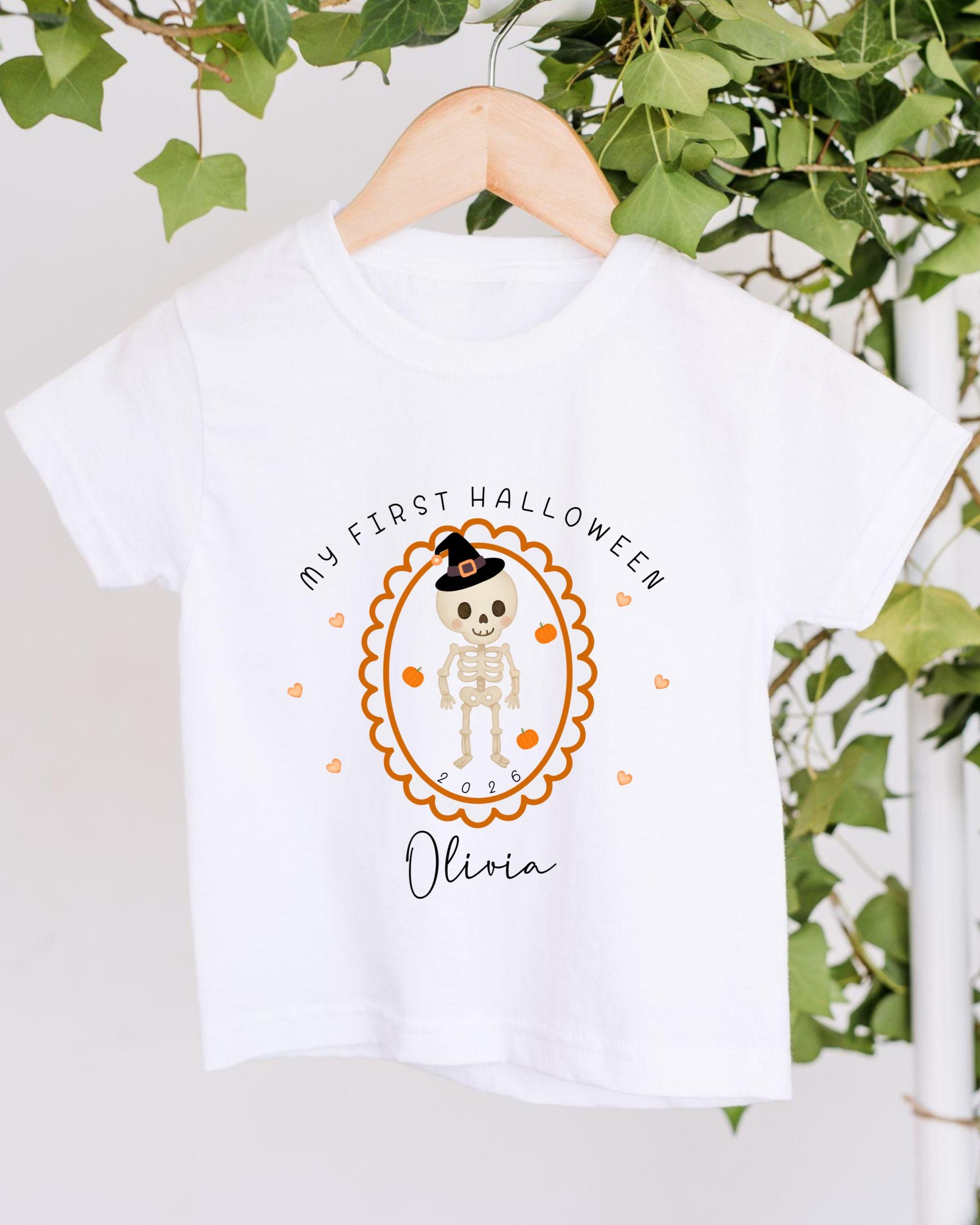 A white t-shirt with a Halloween-themed design hanging on a wooden hanger. The design features a skeleton wearing a witch's hat and surrounded by orange hearts.