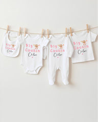 A row of four white baby clothes with the text "Big Cousin Chloe" printed on them, hanging on a clothesline.