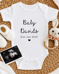 A white baby onesie with the text "Baby Davis Due Jan 2027" printed on it, along with a crocheted bunny toy and a black and white photograph of an infant's head.