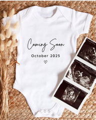A white baby onesie with the text "Coming Soon October 2025" printed on it, along with a heart symbol. The onesie is placed next to three black and white baby ultrasound images.