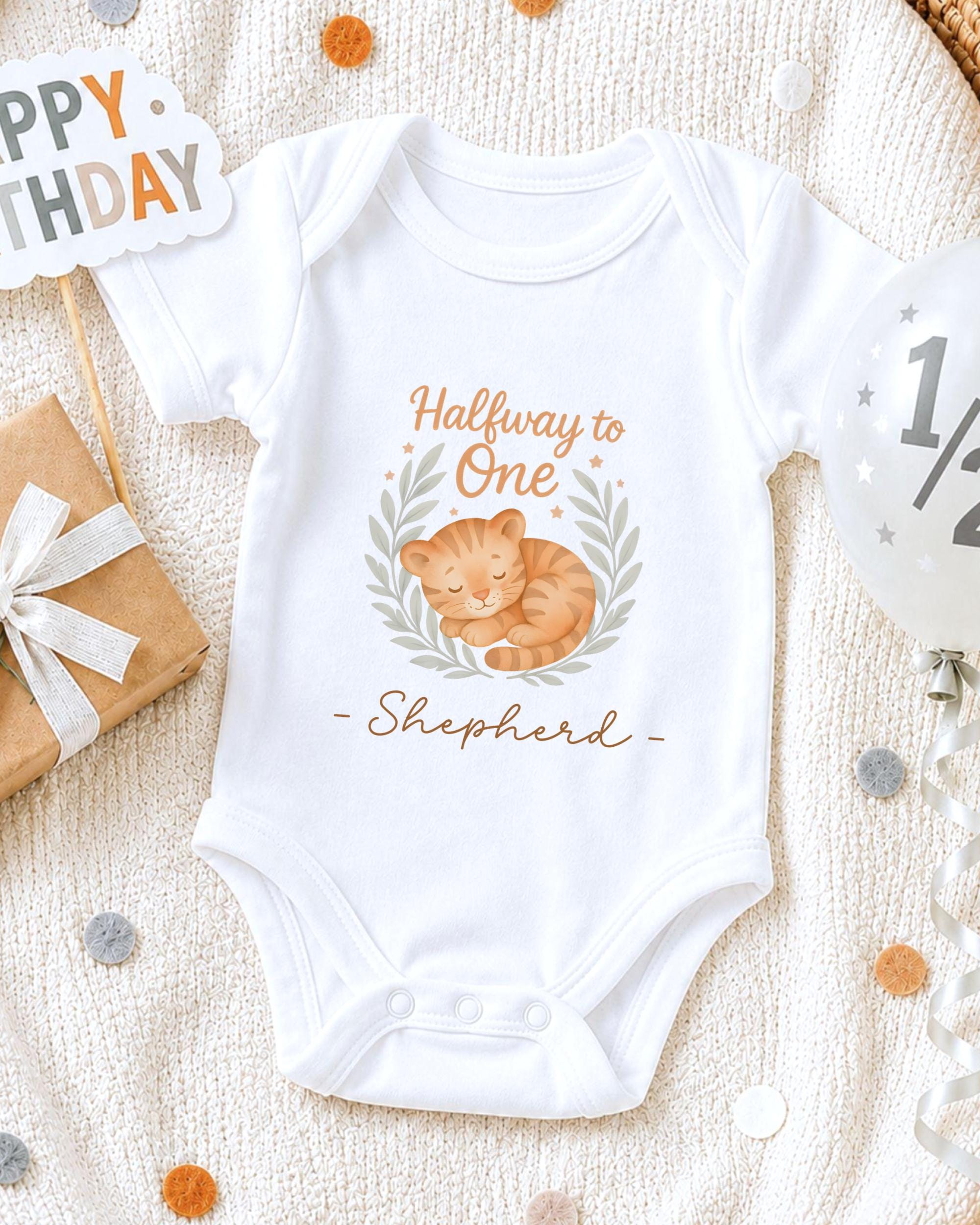 A white onesie with a cat design and the text "Halfway to One - Shepherd" printed on it, along with a "Happy Birthday" sign and confetti.