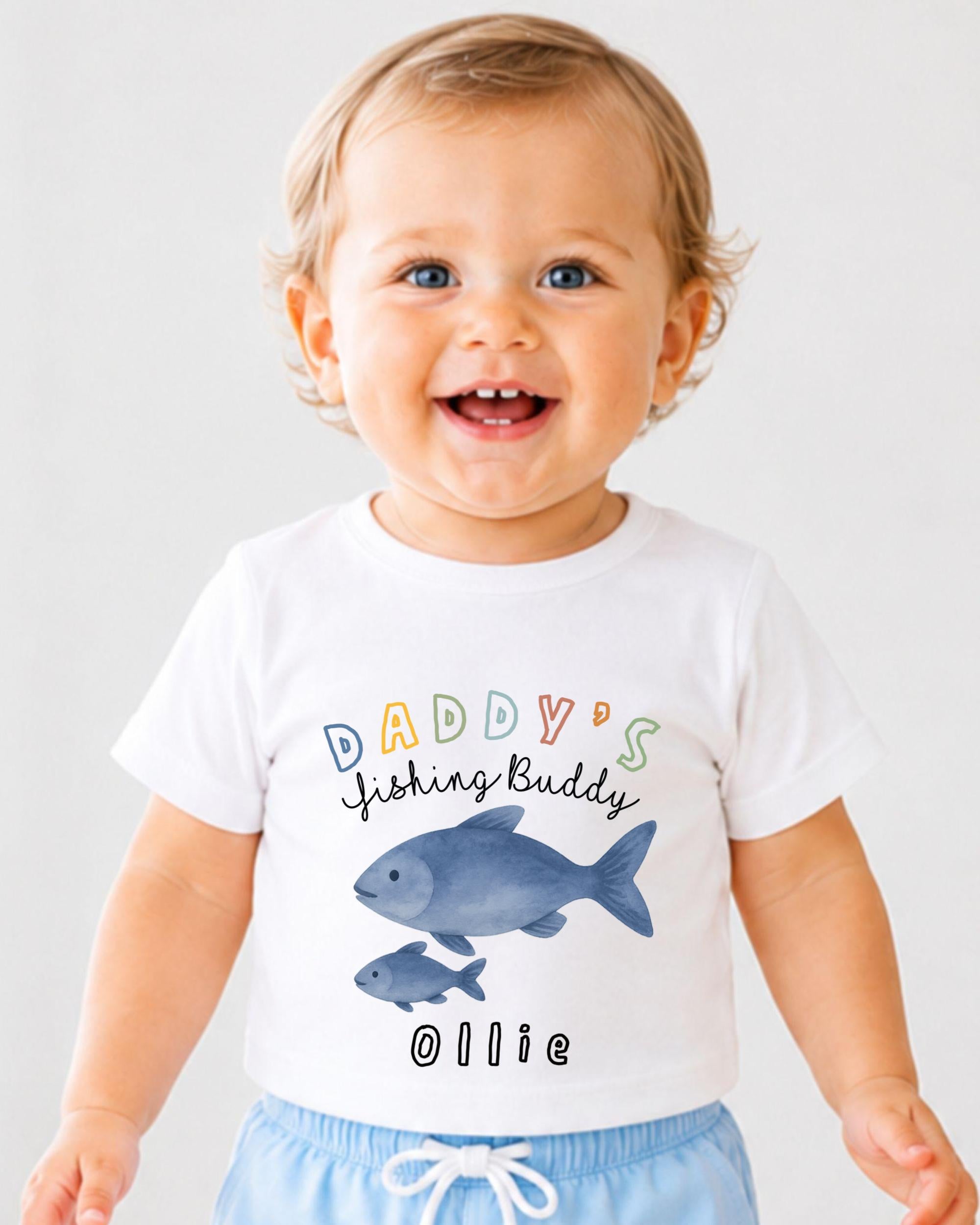 A smiling baby boy wearing a white t-shirt with a fish graphic and text that says "DADDY'S Fishing Buddy Ollie".