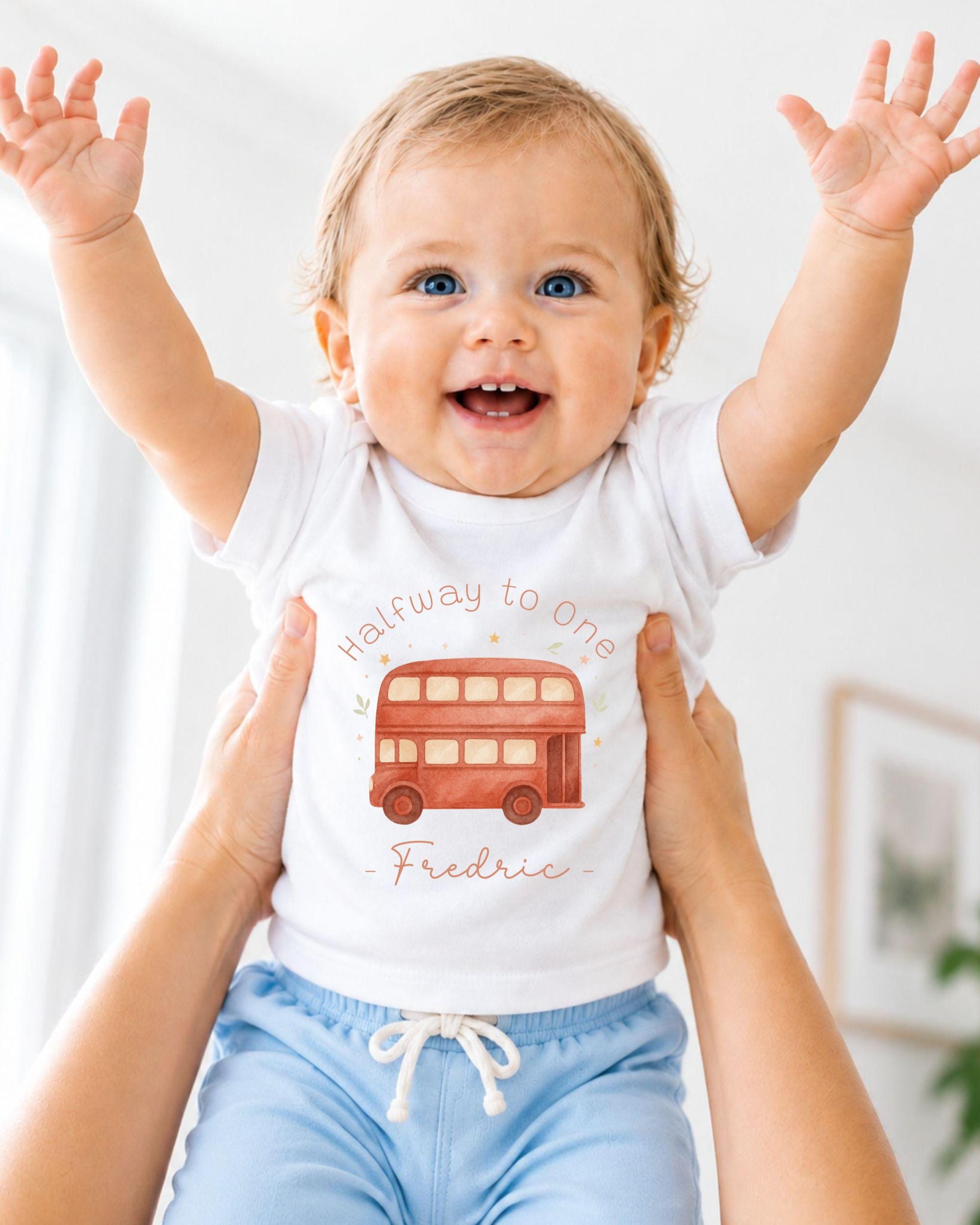 A smiling baby wearing a white t-shirt with a red double-decker bus design and blue shorts, being held up by someone's arms.