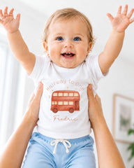 A smiling baby wearing a white t-shirt with a red double-decker bus design and blue shorts, being held up by someone's arms.