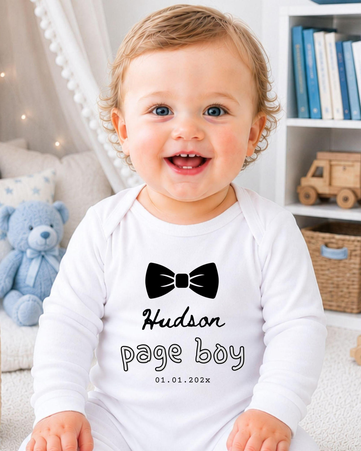 A smiling baby boy wearing a white onesie with a black bow tie and the text "Hudson page boy 01.01.2023" printed on it.