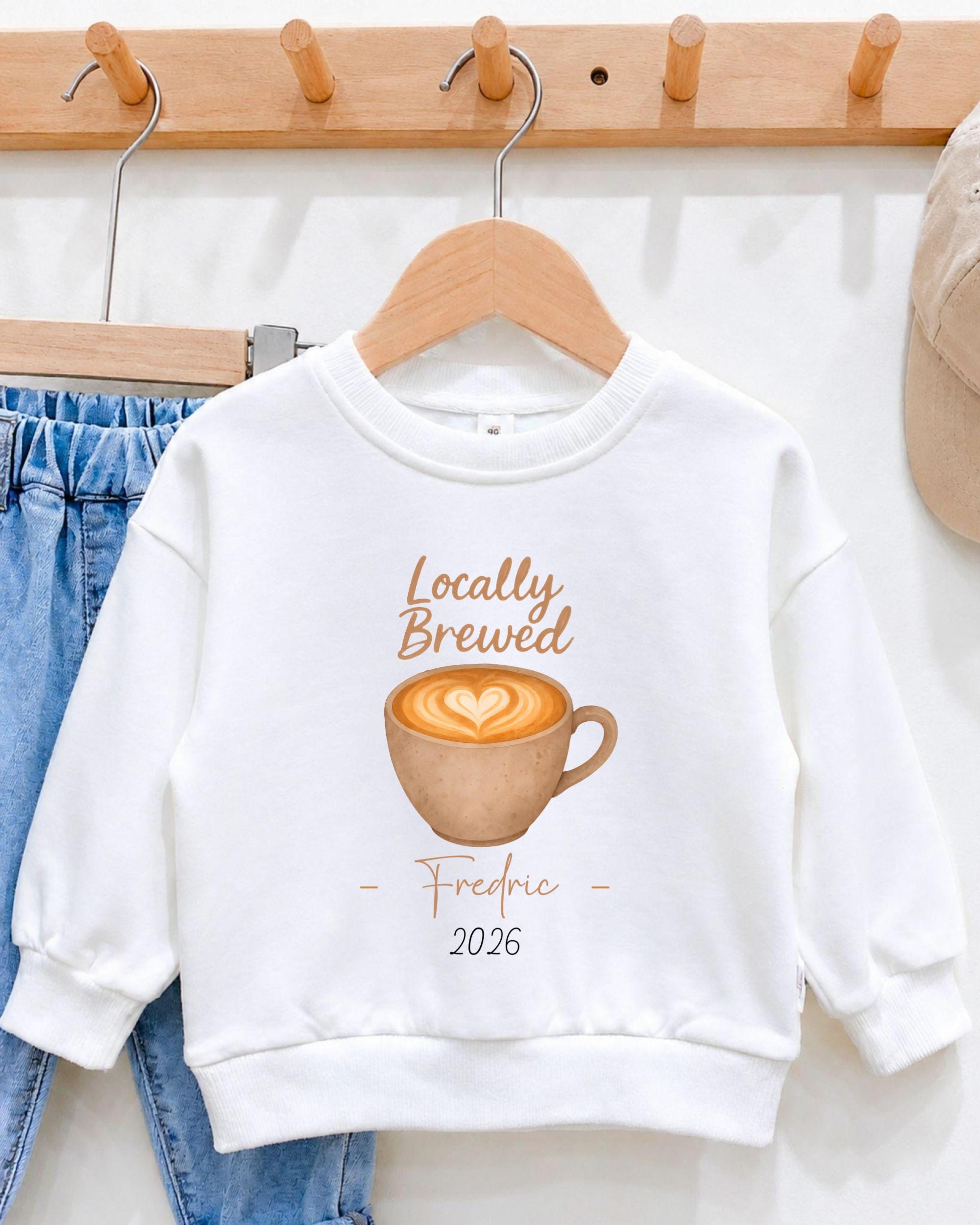 A white sweatshirt with a graphic of a cup of coffee and the text "Locally Brewed - Fredric - 2026" printed on it, hanging on a wooden hanger.