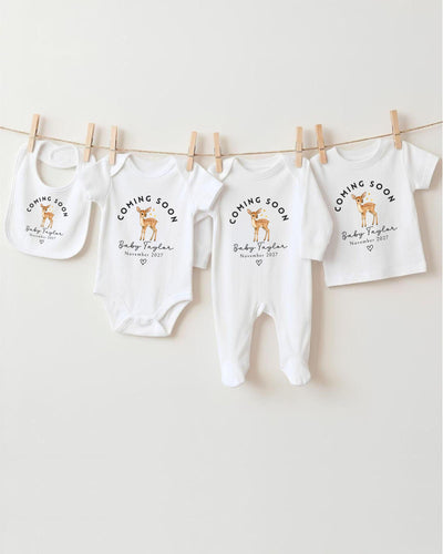 Personalisable white Bib, Baby Vest, Baby Grow and T Shirt with a golden brown watercolour deer beneath curved black 'Coming Soon' lettering, 'Baby Taylor November 2027' text and an outlined heart.