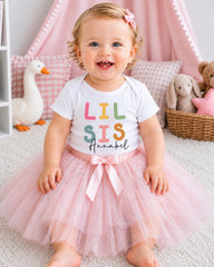 A smiling baby girl wearing a white t-shirt with the text "Lil' sis Annabel" and a pink tutu skirt, sitting on a pink carpet surrounded by stuffed animals.