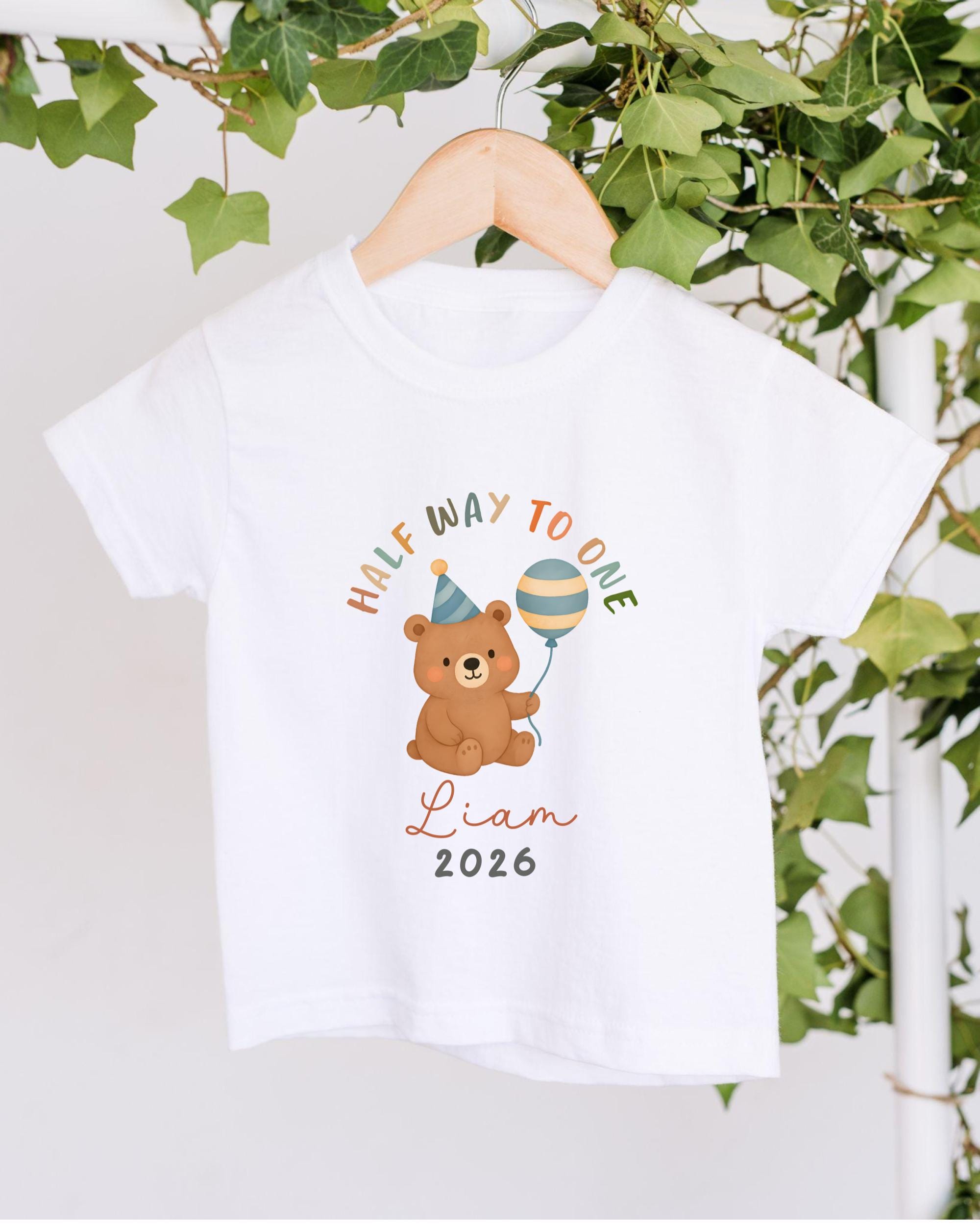 A white t-shirt with a cartoon image of a teddy bear holding a party hat and a balloon, along with the text "Half way to one" and "Liam 2026".