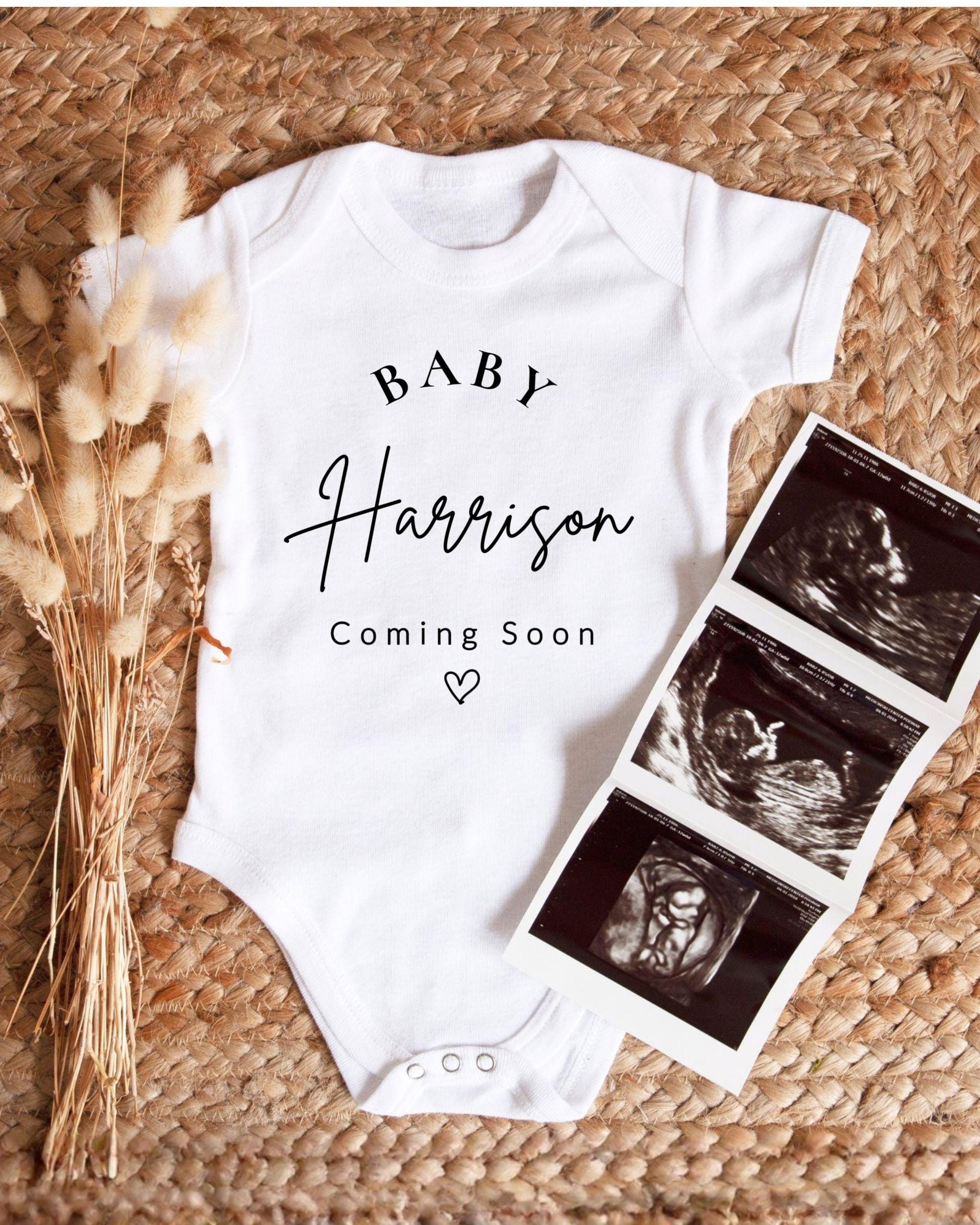 A white baby onesie with the text "BABY Harrison Coming Soon" printed on it, along with a black and white image of an ultrasound of a baby.