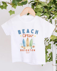 A white t-shirt with a graphic design featuring surfboards and beach elements, along with the text "BEACH CREW BRIGHTON 2027".