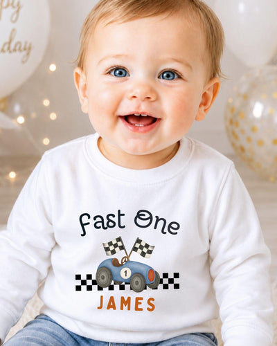 A smiling baby boy wearing a white shirt with a racing car graphic and the text "Fast One James" printed on it.