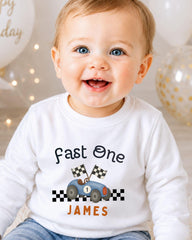 A smiling baby boy wearing a white shirt with a racing car graphic and the text "Fast One James" printed on it.